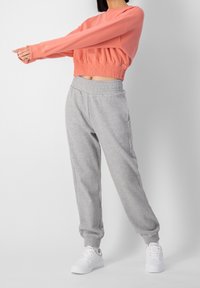 Kvinna som sträcker ut armarna i en korallfärgad kort sweatshirt, gråa sweatpants och vita sneakers mot en helt vit bakgrund.