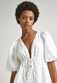Robe blanche avec un décolleté en V profond et des manches bouffantes, ornée de broderies florales et dotée d'une taille à cordon, réalisée en tissu léger.