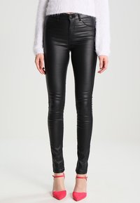 Donna che indossa pantaloni skinny neri in eco-pelle, un maglione bianco peloso e décolleté rosa con cinturino alla caviglia, in piedi di fronte a uno sfondo semplice.