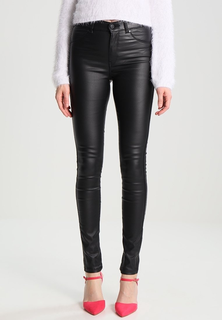 Donna che indossa pantaloni skinny neri in eco-pelle, un maglione bianco peloso e décolleté rosa con cinturino alla caviglia, in piedi di fronte a uno sfondo semplice.