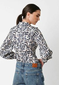 Frau mit einem dunklen Pferdeschwanz, die ein langärmeliges Shirt mit schwarzen und beigen abstrakten Mustern sowie hochgeschnittene blaue Jeans trägt und seitlich schaut.