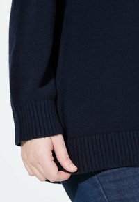 Maglione lavorato a maglia blu navy con polsini e orlo a costine. Texture morbida, vestibilità comoda, mano che afferra delicatamente il bordo inferiore del capo.