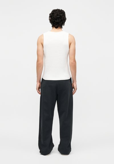 Άτομο με σγουρά μαλλιά που φοράει λευκό αμάνικο τοπ και άνετο μαύρο παντελόνι, στέκεται γυρισμένο από την πλάτη σε απλό φόντο.