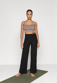 Afskåret brun tanktop og sorte vidde ribbede bukser med en tekstureret overflade, sat mod en simpel hvid baggrund og en grøn yogamåtte.