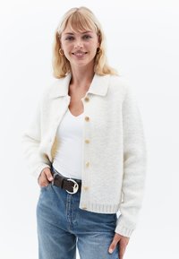 Vit stickad kofta med krage, knappad fram, ribbade muddar och mjuk textur, bärs över en vit topp och blå jeans.