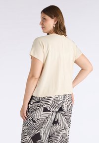 Jasno beżowa bluzka z krótkim rękawem i okrągłym dekoltem, z detalem szwu na plecach, zestawiona z spodniami w czarno-białe liście.