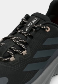 Chaussure de sport noire avec une tige texturée, arborant un logo à trois bandes gris et des lacets à motifs. Conçue avec une semelle coussinée et des oeillets renforcés.