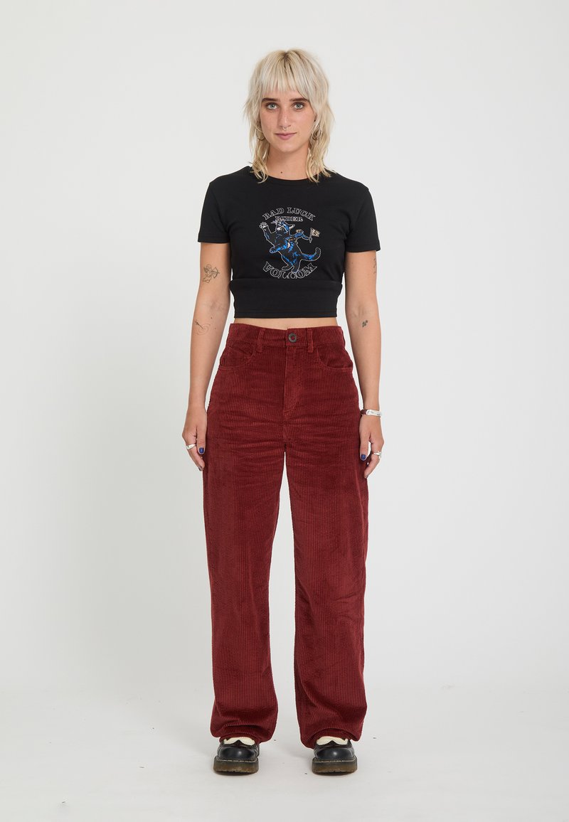 Jeune femme aux cheveux blonds courts portant un crop top graphique noir, un pantalon en velours côtelé rouge taille haute et des chaussures noires, debout sur un fond blanc.