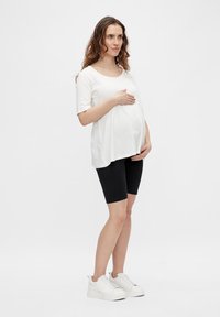 Haut blanc à manches courtes avec un coupe ample sur un ventre de femme enceinte, associé à des shorts de vélo noirs et des baskets blanches. Tissu lisse, design décontracté.