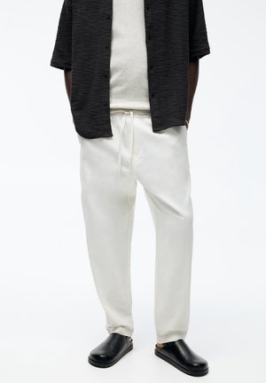 Homme portant un pantalon blanc à cordon, des chaussures noires sans lacets, une chemise beige et une chemise noire texturée à manches courtes boutonnée, les mains dans les poches.