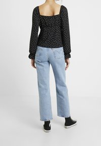 Musta, pitkähiha blouse, jossa on valkoisia pallokuvioita; neliönmuotoinen kaula-aukko ja kerätty tekstuuri. Yhdistetty vaaleansiniisiin suoriin farkkuihin ja mustiin lenkkareihin.