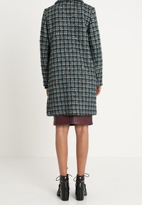 Lång rutig kappa i mörk teal och svart med texturerat tyg. Har en spetsig krage, knäppt fram och en längd som slutar vid mitten av låret.