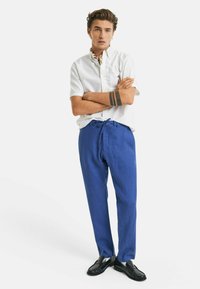 Witte katoenen shirt met korte mouwen, blauwe linnen broek met een trekkoord en zwarte instappers. Model staat met de armen gekruist voor een witte achtergrond.