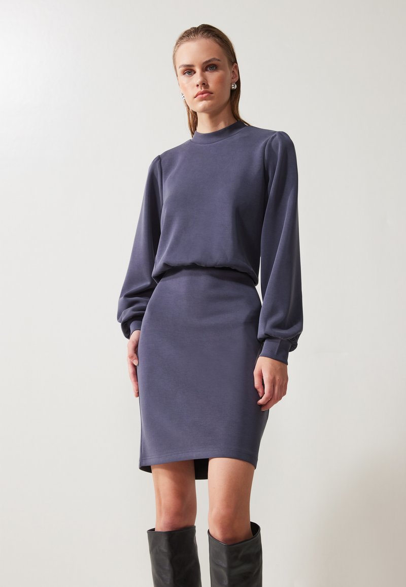 Robe longue violette à manches longues avec une taille cintrée et un ourlet droit, fabriquée en tissu doux. Les détails incluent des manches bouffantes et un col haut.