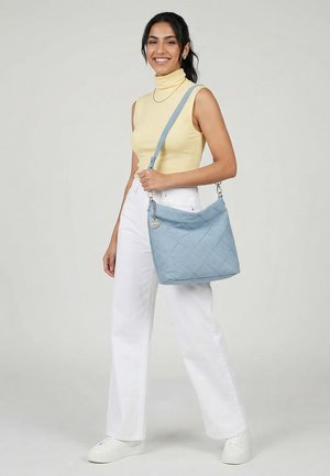 Naeratav tumedate juustega naine kollase varrukateta kõrgkaelusega pluusi ja valgete pükstega, kandes sinist pitseeritud õlakotti.