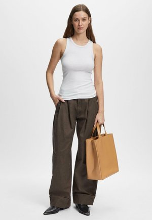 Frau trägt weißes ärmelloses Oberteil und dunkle, weit geschnittene Hose, hält eine beige Tragetasche und trägt schwarze spitze Schuhe, steht vor einem schlichten Hintergrund.