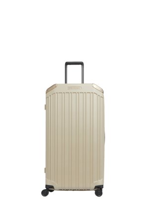 Beige harde koffer met vier wielen, verticale groeven, metalen hoekaccenten en een uitgeschoven zwart handvat, getoond tegen een witte achtergrond.