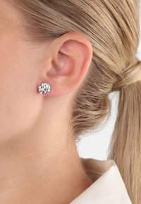 Silverfärgade trädformade örhängen med klara rhinestones, med en strukturerad yta, visade på en närbild av en models örat.