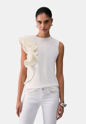 Donna che indossa un top bianco senza maniche con un grande dettaglio a balza sulla spalla sinistra, abbinato a pantaloni bianchi e orecchini a cerchio argentati con braccialetti.