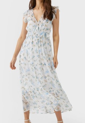Mujer con vestido blanco sin mangas con estampado floral en azul y verde y detalles fruncidos en los hombros, posando sobre un fondo liso.