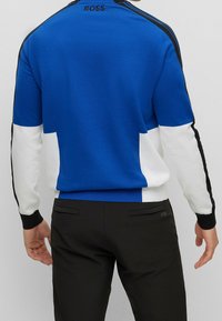 Hombre vistiendo un suéter de manga larga azul, blanco y negro con el texto "BOSS", combinado con pantalones negros, visto desde atrás.