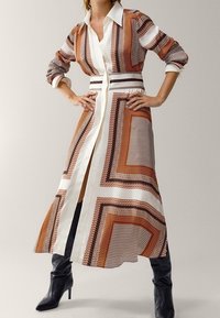 Robe chemise longue avec un motif géométrique marron et blanc, devant à boutons, et ceinture. Associée à des bottes noires arrivant au genou.