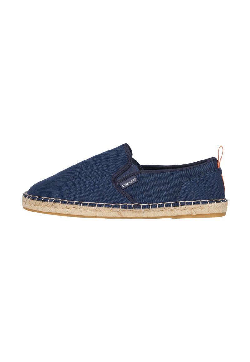 Zapato de lona azul marino tipo slip-on con suela de cuerda de yute, que presenta un borde cosido y una pequeña etiqueta de logo en el lateral. Sutil acento en color naranja en el talón.