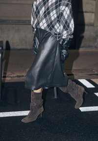 Pull en argyle noir et blanc, jupe en cuir noire, bottes à talons souples en daim marron. Textures brillantes et structurées avec un design superposé.