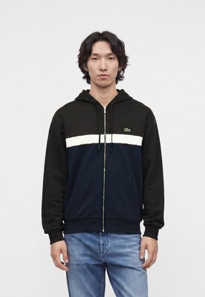 Mann mit schwarzem und marineblauem Zip-up-Hoodie mit weißem Streifen und Logo, trägt hellblaue Jeans, steht vor einfarbig hellem Hintergrund.