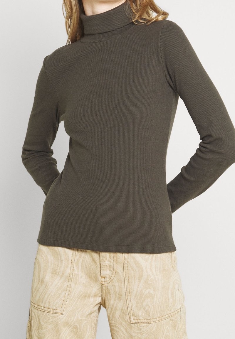 Personne portant un haut moulant à col roulé manches longues couleur olive foncé et un pantalon beige avec un motif ondulé subtil, mains posées sur les hanches.