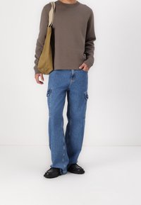 Brauner Rundhals-Pullover, hellblaue Cargo-Jeans mit Seitentaschen und schwarze Schuhe. Das Modell hält eine beigefarbene Tote-Tasche mit einem Schulterriemen.