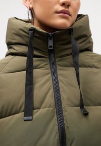 Giacca puffer verde oliva con colletto alto, chiusura frontale con cerniera e lacci neri. Presenta una trama trapuntata e accessori con marchio.