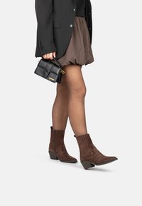 Botas de tobillo de ante marrón con punta afilada y tacón curvado. Presentan detalles acanalados verticales y una cremallera lateral. Combinadas con una falda marrón.