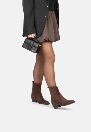 Botas de tobillo de ante marrón con punta afilada y tacón curvado. Presentan detalles acanalados verticales y una cremallera lateral. Combinadas con una falda marrón.