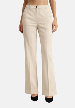Pantaloni eleganti beige con piega frontale, gambe dritte, chiusura con bottone e cerniera, indossati con scarpe nere con punta appuntita.