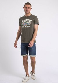 T-shirt en coton vert olive avec un graphisme "HONOLULU", associé à un short en denim bleu foncé. Des baskets blanches complètent la tenue décontractée.