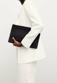 Henkilö valkoisessa puvussa pitää kädessään teksturoitua mustaa kudottua laukkua vaaleaa taustaa vasten.