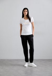 T-shirt bianca a maniche corte con logo "EA7" in oro, abbinata a pantaloni in felpa neri con logo ricamato, completata da sneakers bianche.