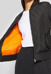 Frau trägt schwarze gesteppte Bomberjacke mit leuchtend orangefarbenem Futter, weißes, kurz geschnittenes T-Shirt und schwarze Hosen mit elastischem Bund.