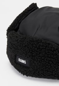 Chapéu preto com uma aba suave e texturizada parecida com pelo e um topo liso e brilhante. Apresenta um rótulo retangular com a marca "RAINS."