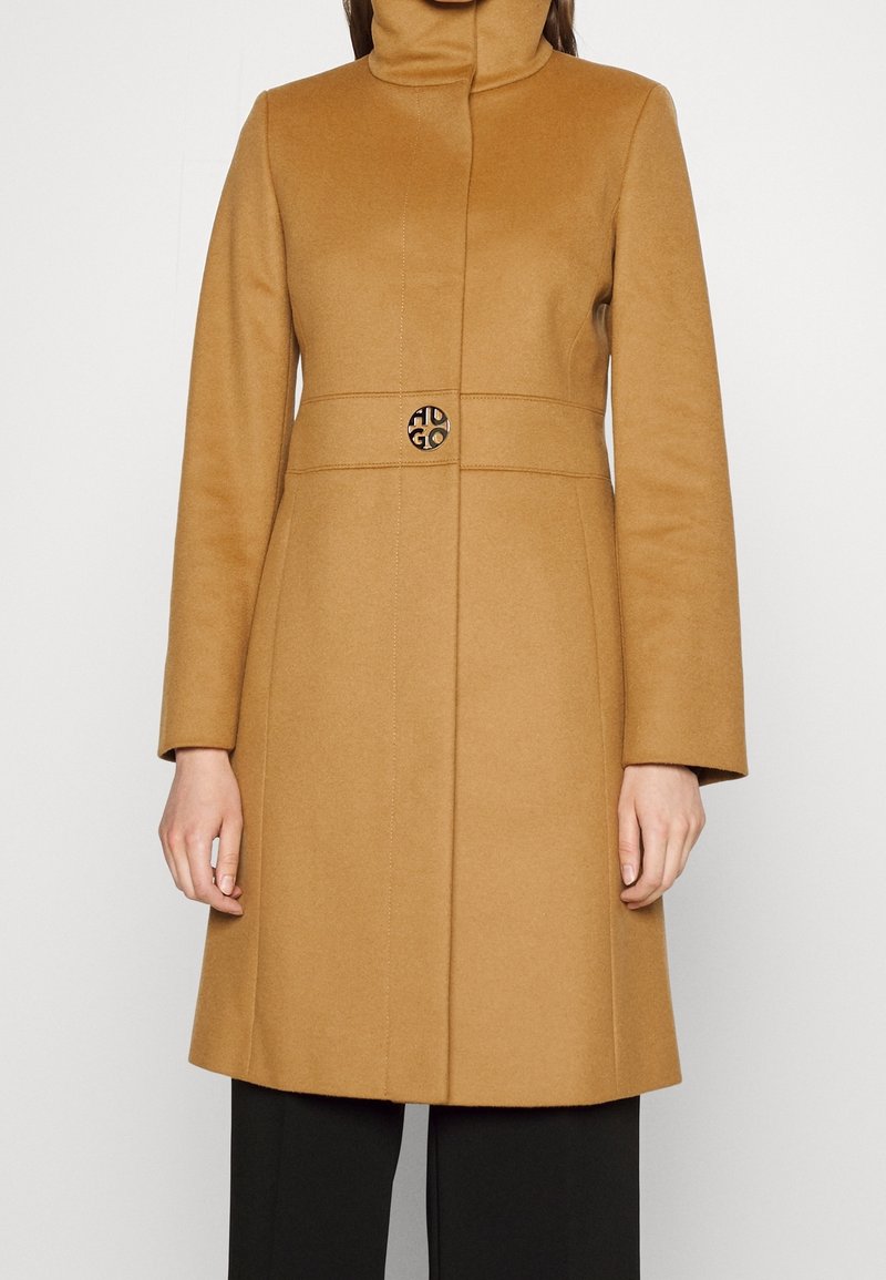 Femme portant un manteau couleur camel, longueur genou, avec un col haut et un bouton décoratif unique à la taille, associé à un pantalon noir.