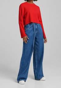 Osoba ubrana w luźną czerwoną bluzkę z długim rękawem, wysokowysokie szerokie niebieskie dżinsy i białe trampki, stojąca na tle szarego tła.