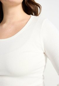 Maglione bianco a maniche lunghe con scollatura a barca, caratterizzato da una texture morbida e un design aderente che mette in risalto l'elasticità del tessuto.