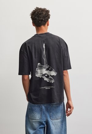 Άτομο με σγουρά μαλλιά που φοράει μαύρο t-shirt με γραφικό σχέδιο κρεμασμένων ποδοσφαιρικών παπουτσιών στην πλάτη και φαρδιά μπλε τζιν παντελόνια.