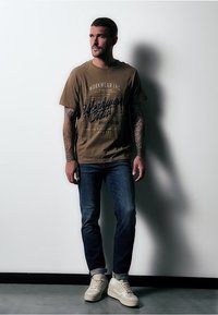 Hnědý grafický tričko s nápisem "WORKWEAR INC", modré džíny a béžové tenisky. Model stojí u jednoduché zdi, ruce uvolněné.