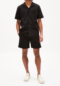 Czarna koszula z krótkim rękawem i kieszenią na piersi, zestawiona z czarnymi szortami z podwiniętym brzegiem. Model nosi białe buty wsuwane.