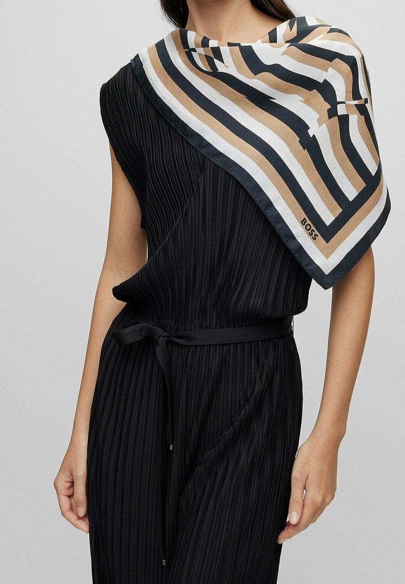 Femme portant une robe noire plissée sans manches avec une ceinture noire et une écharpe beige, noire et blanche à rayures drapée sur une épaule.