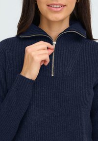 Sötétkék bordázott, zipzáros pulóver magas gallérral és fém zipzár részletekkel. Az anyaga puha és kötött, szoros illeszkedésű.