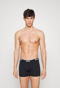 Schwarze Boxershorts mit einem weißen Logo-Bund. Der Stoff wirkt glatt und dehnbar und ist für Komfort und Unterstützung ausgelegt.