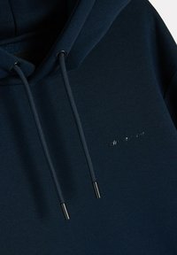 Granatowy hoodie wykonany z miękkiego materiału, z kapturem na sznurki, czarnymi metalowymi końcówkami sznurków oraz małym metalicznym logo z przodu.